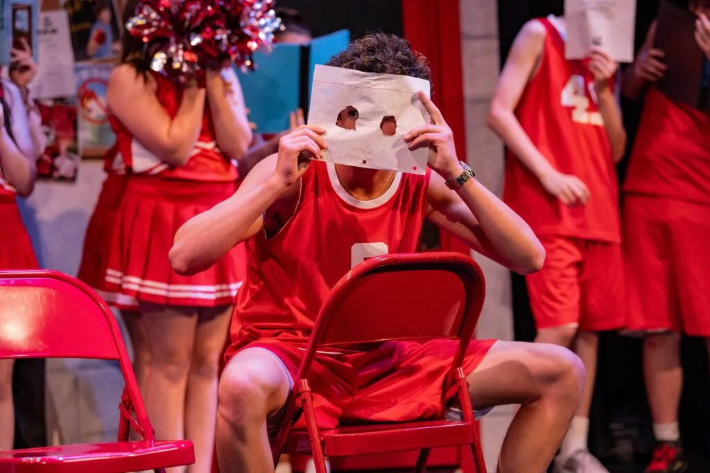 A cast member in a red basketball jersey holds a sheet of paper with eye holes over his face while other cast members stand behind him, in a Ritz Theatre Company youth production. Photo: Zachary Moore, courtesy of The Ritz Theatre Company