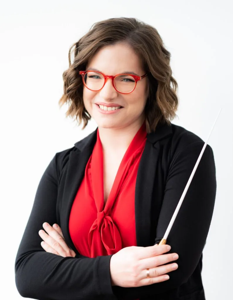 Noam Aviel, music director of Symphony in C, smiling in a red blouse and black jacket and holding a conductor baton. Photo: Siggi Ragnar